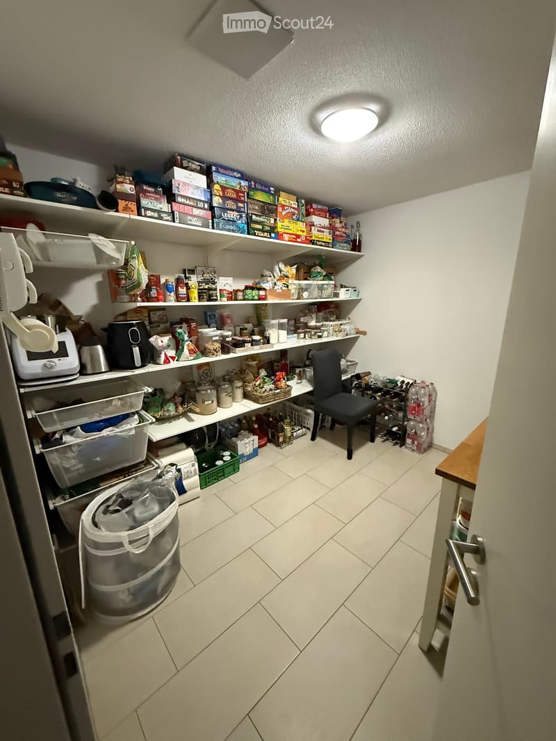 Well-organized pantry with multiple shelves filled with food items, board games, and kitchen appliances. Includes a small black chair, a trash bin, and a wine rack.
