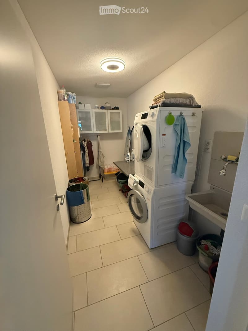 A small, well-lit laundry room with tiled flooring, a washing machine, dryer, sink, and various storage items.