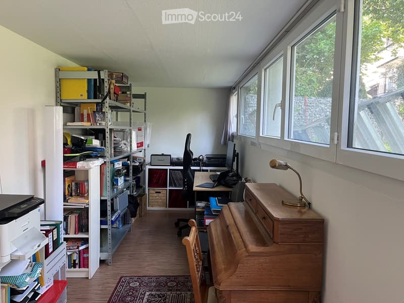 A well-lit study or office space with wooden flooring, large windows, a desk with a laptop, a bookshelf filled with books and office supplies, a printer, and a small table lamp on a wooden cabinet.