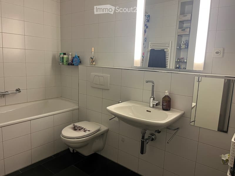 Bathroom with white tiled walls, a sink with a modern faucet, a toilet, a bathtub, a mirror, and a towel on a rack. Shelves hold various toiletries and a decorative bottle.