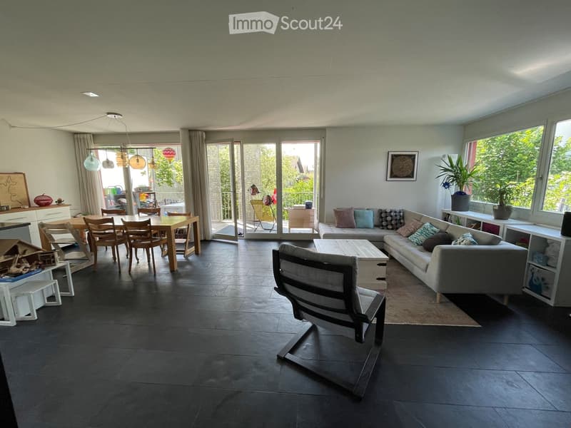 Open-plan living and dining area with dark tiled flooring, large windows, and a balcony access. Features a grey sectional sofa, coffee table, dining table with chairs, pendant lights, and shelves with decorative items.