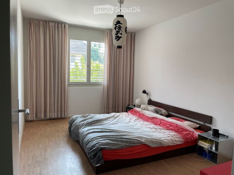 A furnished bedroom with a wooden bed, red and grey bedding, a nightstand with a lamp, and striped curtains on the windows.