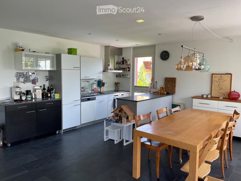 Modern kitchen with white cabinets, dark countertops, and a wooden dining table with chairs. Features include a refrigerator, dishwasher, coffee maker, and various kitchen appliances.