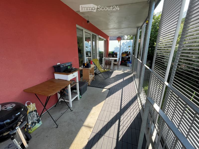 Balcony with tiled floor, wooden table, grill, and chairs, adjacent to a red wall and open sliding doors leading to an indoor area.