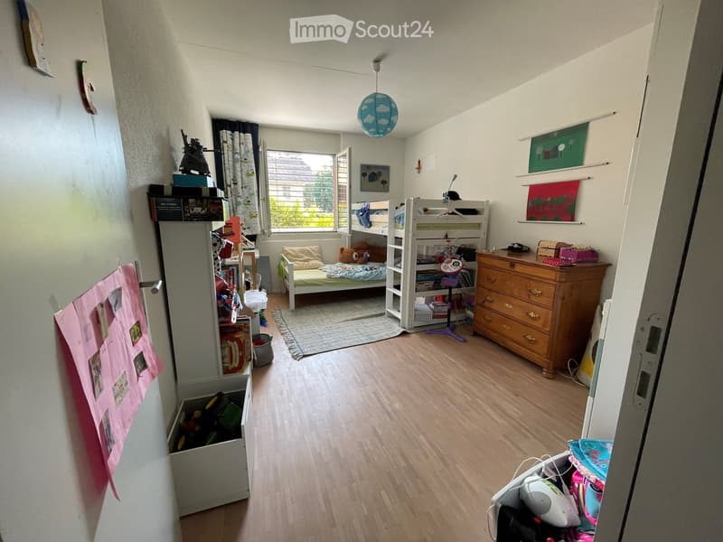 A cozy bedroom with wooden flooring, a bunk bed, a bookshelf, a wooden dresser, and various toys and decorations.