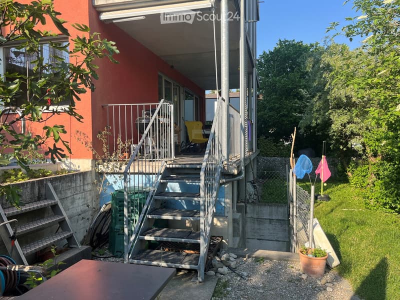 Outdoor area with a staircase leading to a balcony, a small garden with potted plants, and a grassy area.