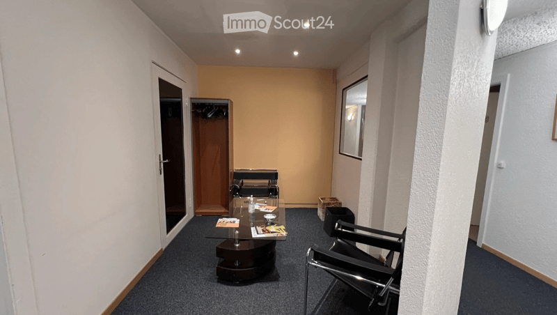 Empty room with carpeted floor, glass table, modern chairs, cabinet with clothes hanging, trash can, door on the right, window, mirror
