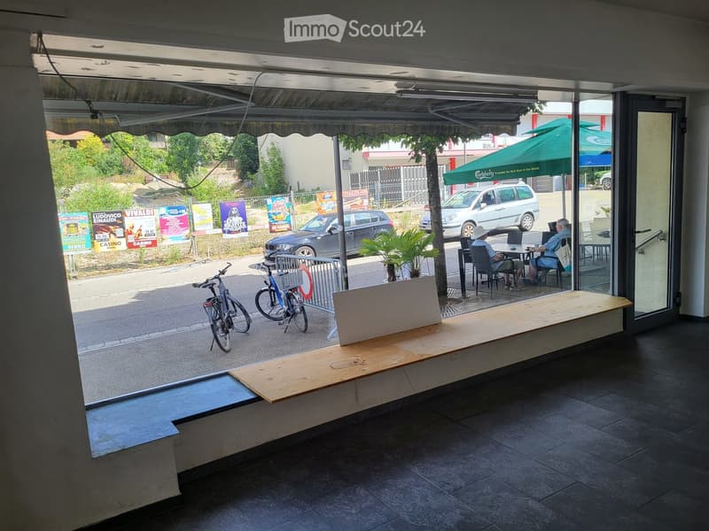 View from inside a building looking out to a covered outdoor seating area with tables and chairs, bicycles parked nearby, and a green umbrella visible outside.