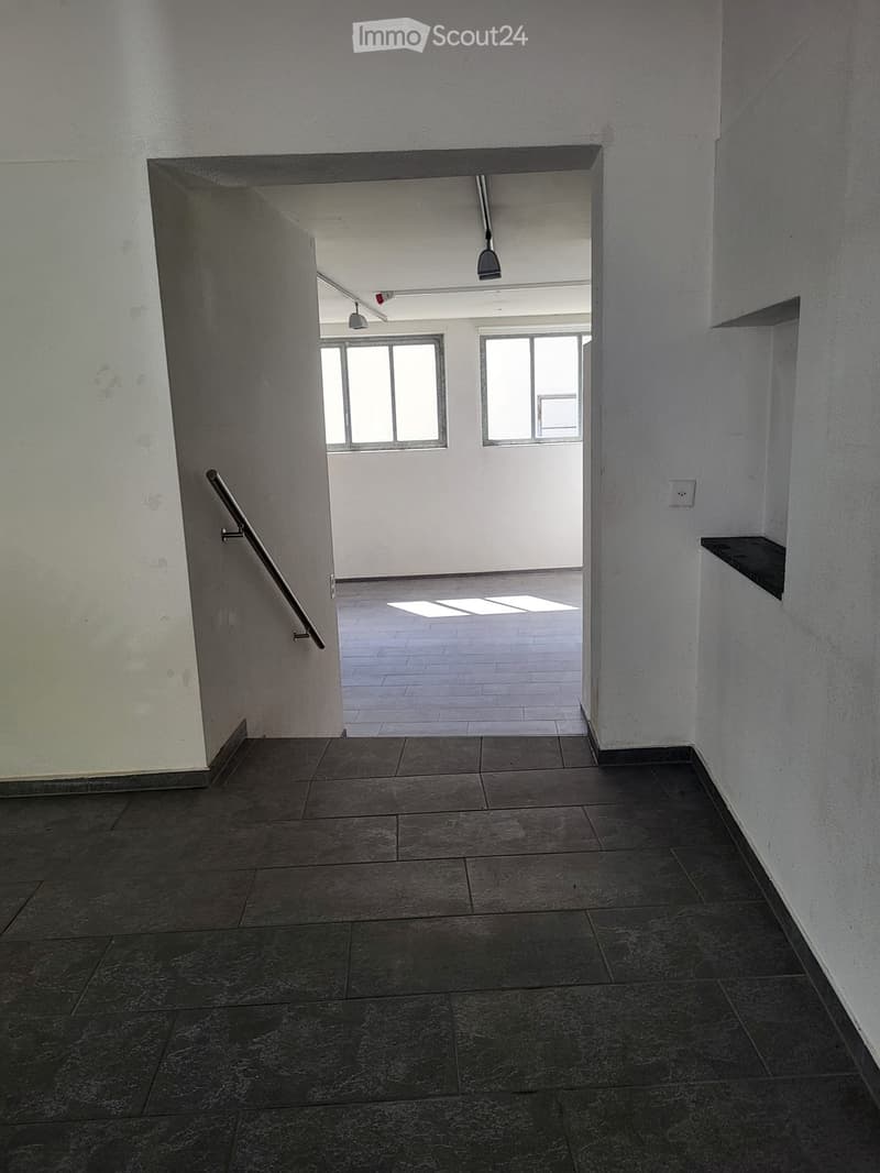 Empty hallway with tiled floors, white walls, and a metal handrail leading to an open area with windows and a ceiling light fixture.