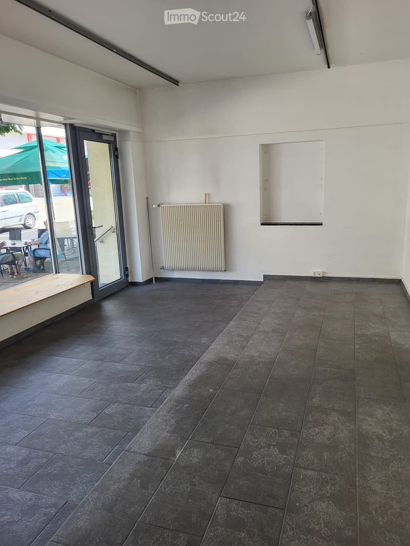Empty room with dark tiled flooring, white walls, a radiator, and a large glass door leading outside. A small wooden bench is visible near the door.