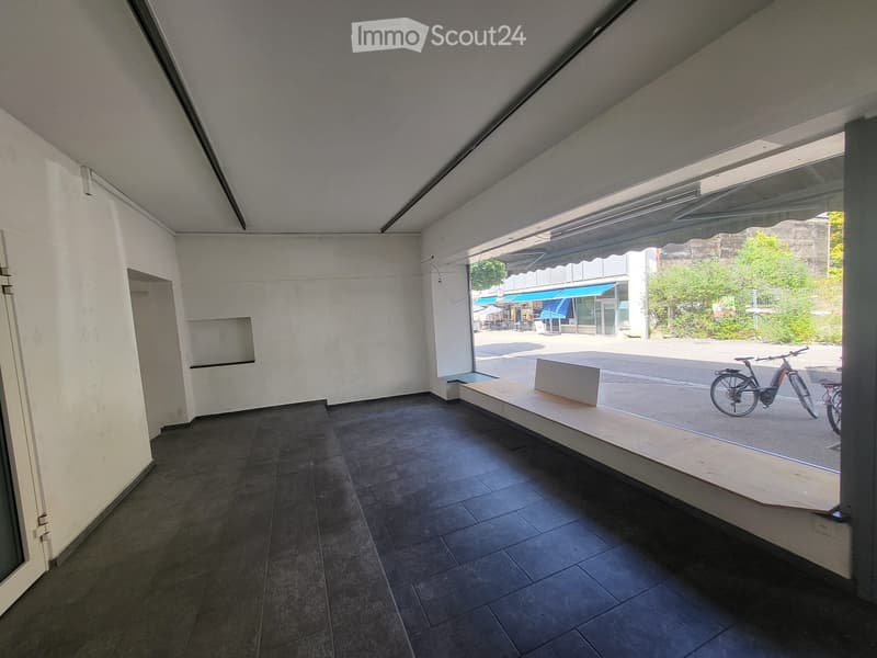 Empty room with dark tiled floor, white walls, and large glass window opening to an outdoor area with bicycles and a bench.
