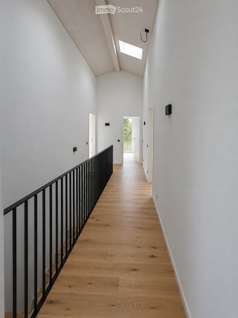 Modern hallway with light wooden flooring, white walls, and a black metal railing leading to a bright room with a view of greenery.