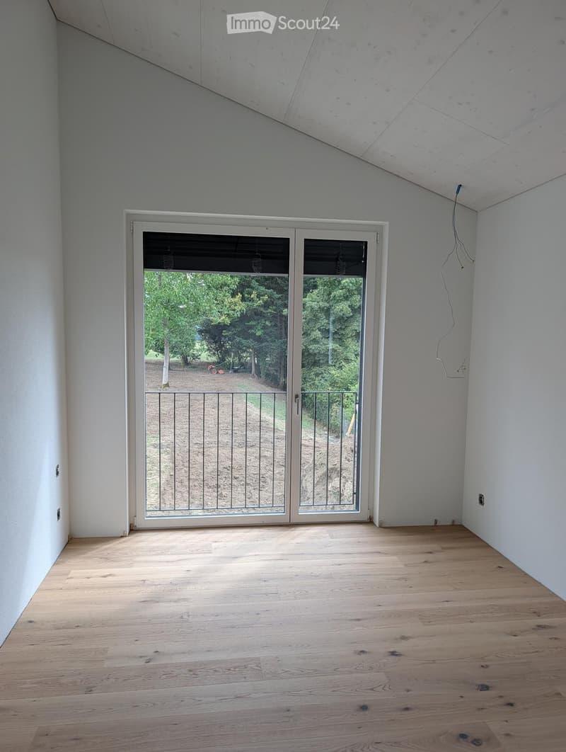 Empty room with light wooden flooring, white walls, and a large sliding glass door leading to an outdoor area with trees and a gravel path.