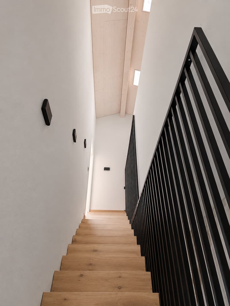 Modern staircase with wooden steps and a sleek black metal railing, set against white walls and a light-colored ceiling.
