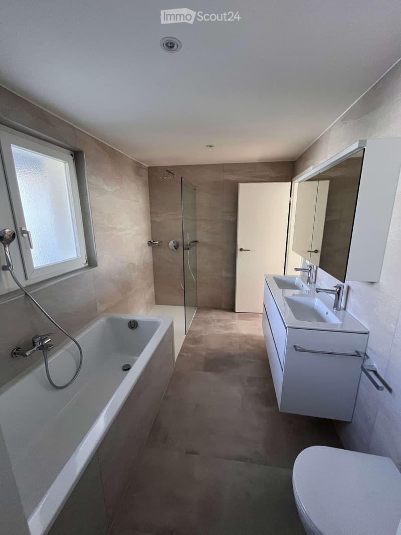 Modern bathroom with a bathtub, double sink, toilet, shower, and large mirror. Features a window and neutral-colored tiles.