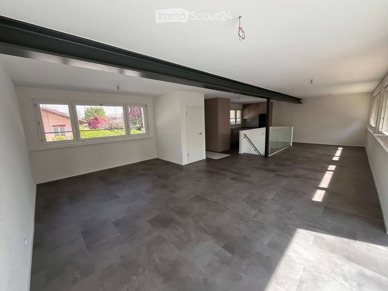 Empty room with dark wooden flooring, white walls, large windows, and a visible kitchen area in the background.