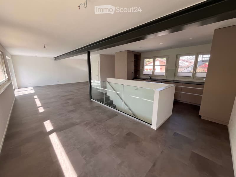 Open-plan space with modern interior design, featuring a glass staircase, large windows, and a kitchen area with cabinets and a sink.