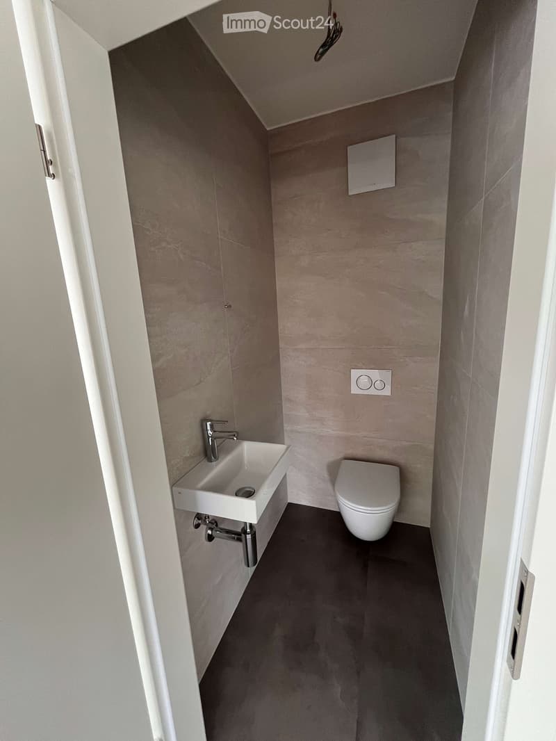Modern bathroom with a white sink, chrome faucet, and white toilet. Beige tiled walls and dark grey floor.