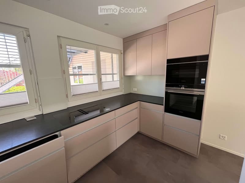 Modern kitchen with light-colored cabinets, black countertop, and built-in oven. Windows with blinds provide natural light.