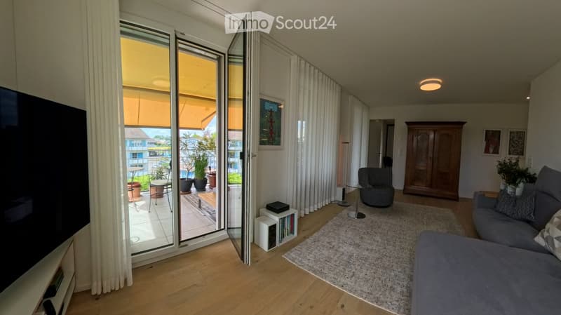 Modern living room with large sliding glass doors leading to a balcony, grey sectional sofa, coffee table, armchair, cabinet, and potted plants.
