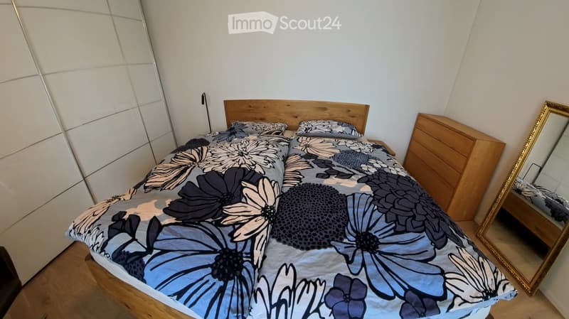 A modern bedroom with a wooden bed frame, a floral-patterned bedspread, matching pillows, a large mirror, and a wooden wardrobe.