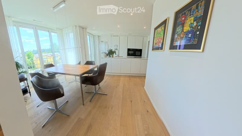 Bright, modern room with light wooden flooring, large windows, a dining table with chairs, white cabinets, and framed artwork on the walls.