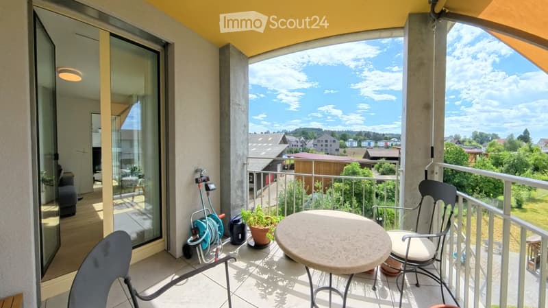 Balcony with a round table, two chairs, and a potted plant, overlooking a suburban landscape with houses and greenery.