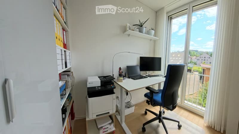 A modern, well-lit home office with a white desk, ergonomic chair, laptop, monitor, printer, and shelves filled with binders and books. Large windows offer a view of the outdoor area.