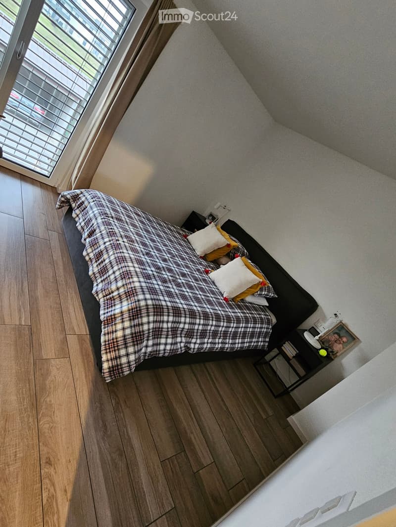 Cozy bedroom with a wooden floor, a bed with a plaid duvet and pillows, a nightstand with a lamp, and a picture frame on top.