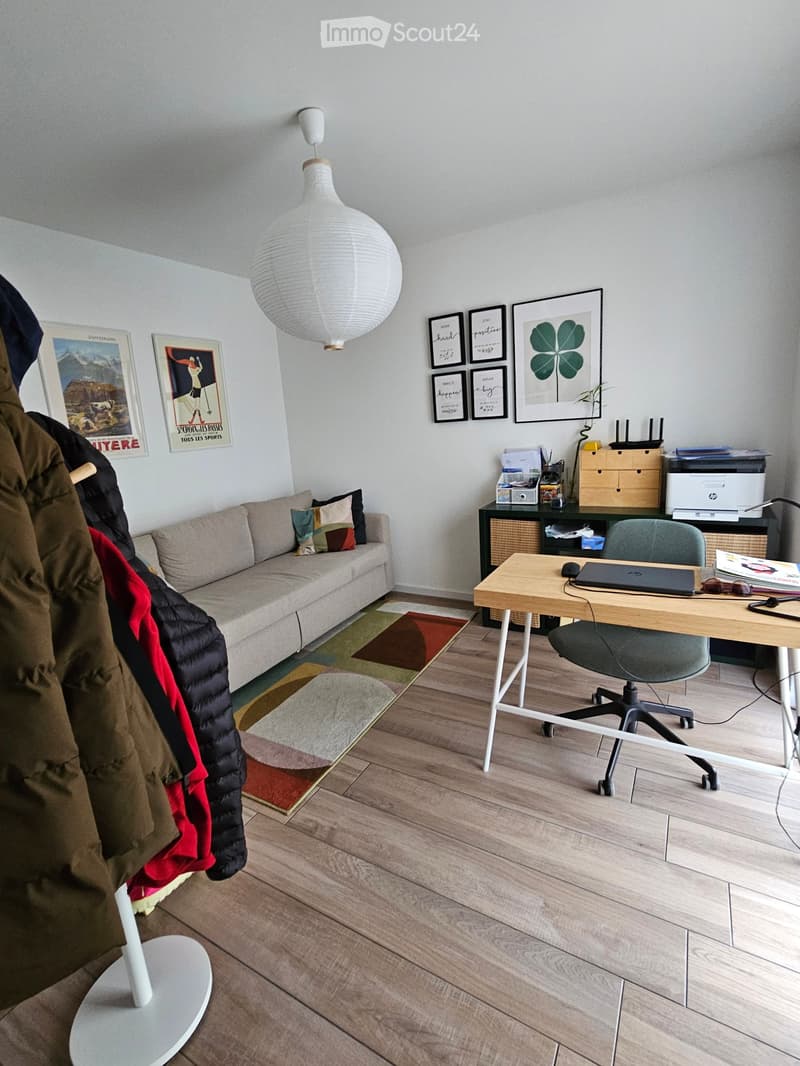 A modern, well-lit room with a sofa, a desk with a laptop, a hanging lamp, and several framed pictures on the wall. The room features wooden flooring and a colorful rug.