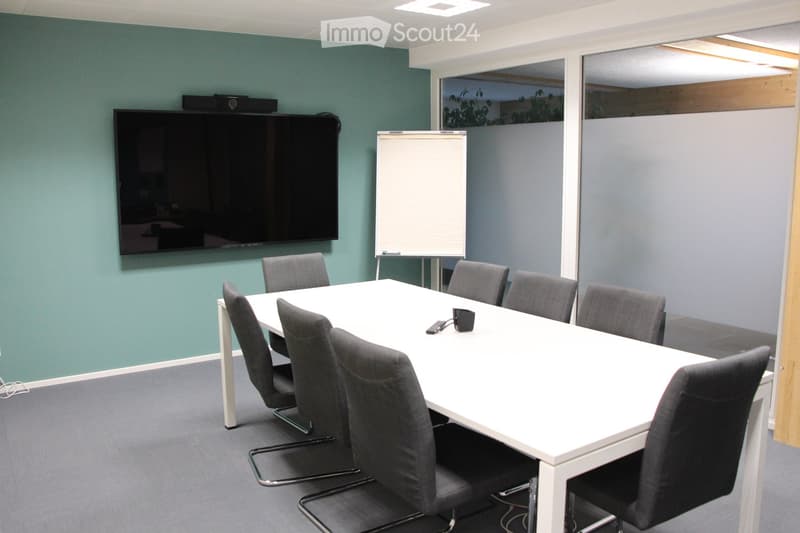 A modern conference room with a white rectangular table surrounded by grey chairs, a large flat-screen TV mounted on the wall, and a whiteboard on a stand.