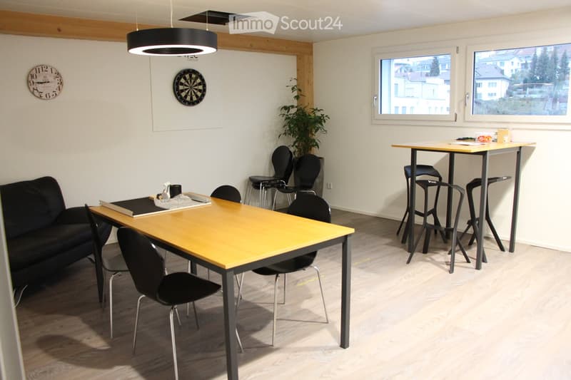 Bright room with a sofa, two tables with chairs, a dartboard, a clock, and a plant. Large windows provide natural light.