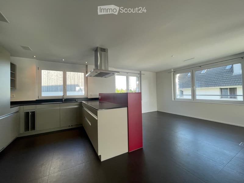 Modern kitchen with dark flooring, white cabinets, a red accent wall, and a central island with a sink and range hood. Large windows provide natural light.