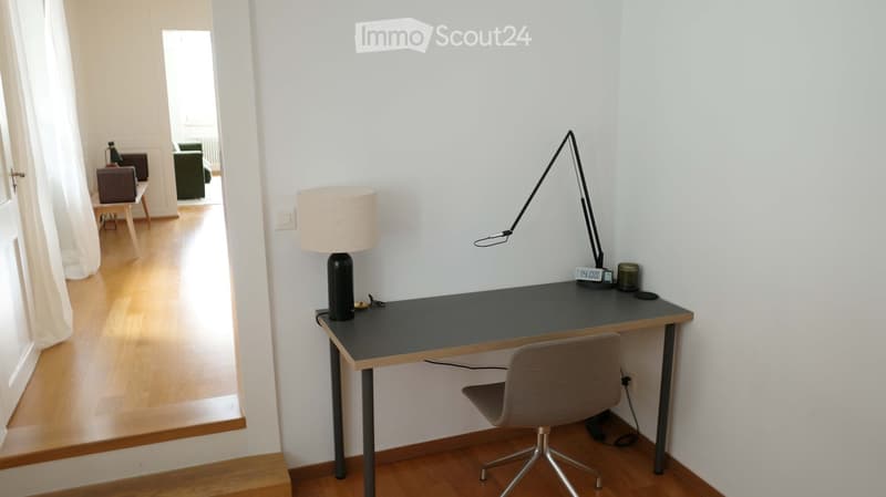 A modern, minimalist room with a wooden floor, a black desk, a grey chair, a table lamp, and a floor lamp. The room has white walls and a view into another room with a green armchair and a small table.