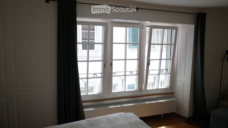 Bedroom with wooden floor, large window with white frame and curtains, radiator below the window, bed visible in the foreground.