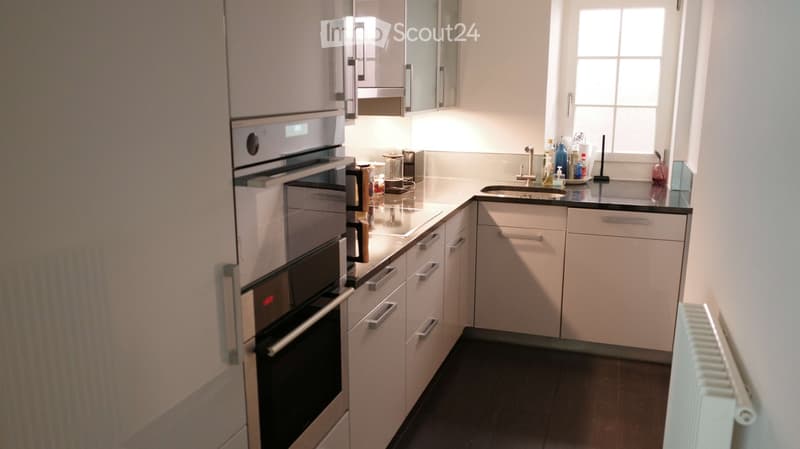 Modern kitchen with white cabinets, stainless steel oven, sink, and various appliances. Features a window above the sink and a radiator on the wall.
