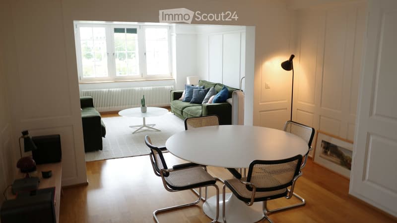 Bright living and dining area with a green sofa, white coffee table, dining table with chairs, and a floor lamp. The space features large windows and wooden flooring.