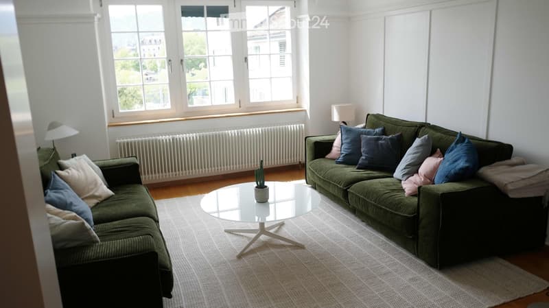 A modern living room with a green sectional sofa, a round glass coffee table, and a small potted plant. The room features large windows, a radiator, and a neutral color palette.