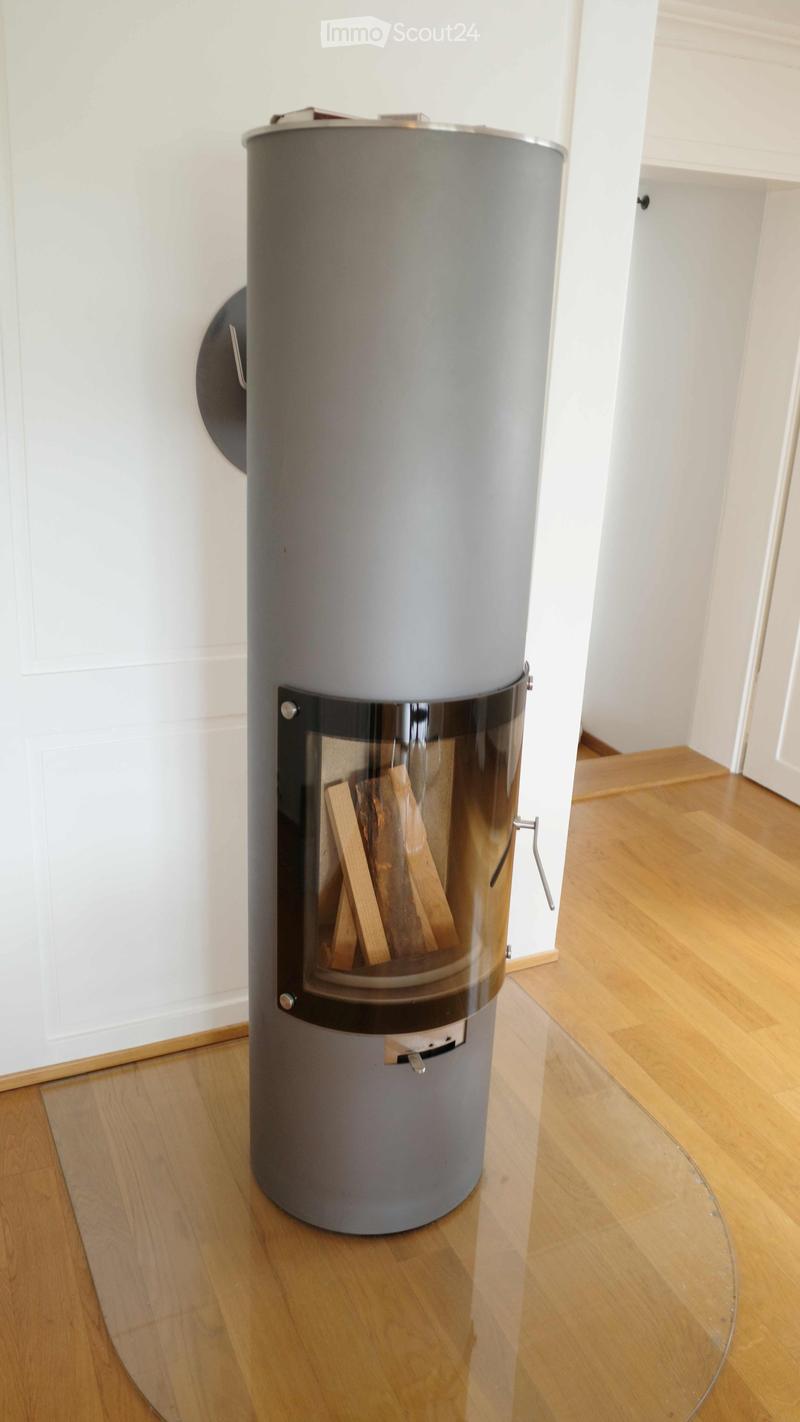 Modern cylindrical fireplace with glass front, wood logs inside, placed on wooden floor.