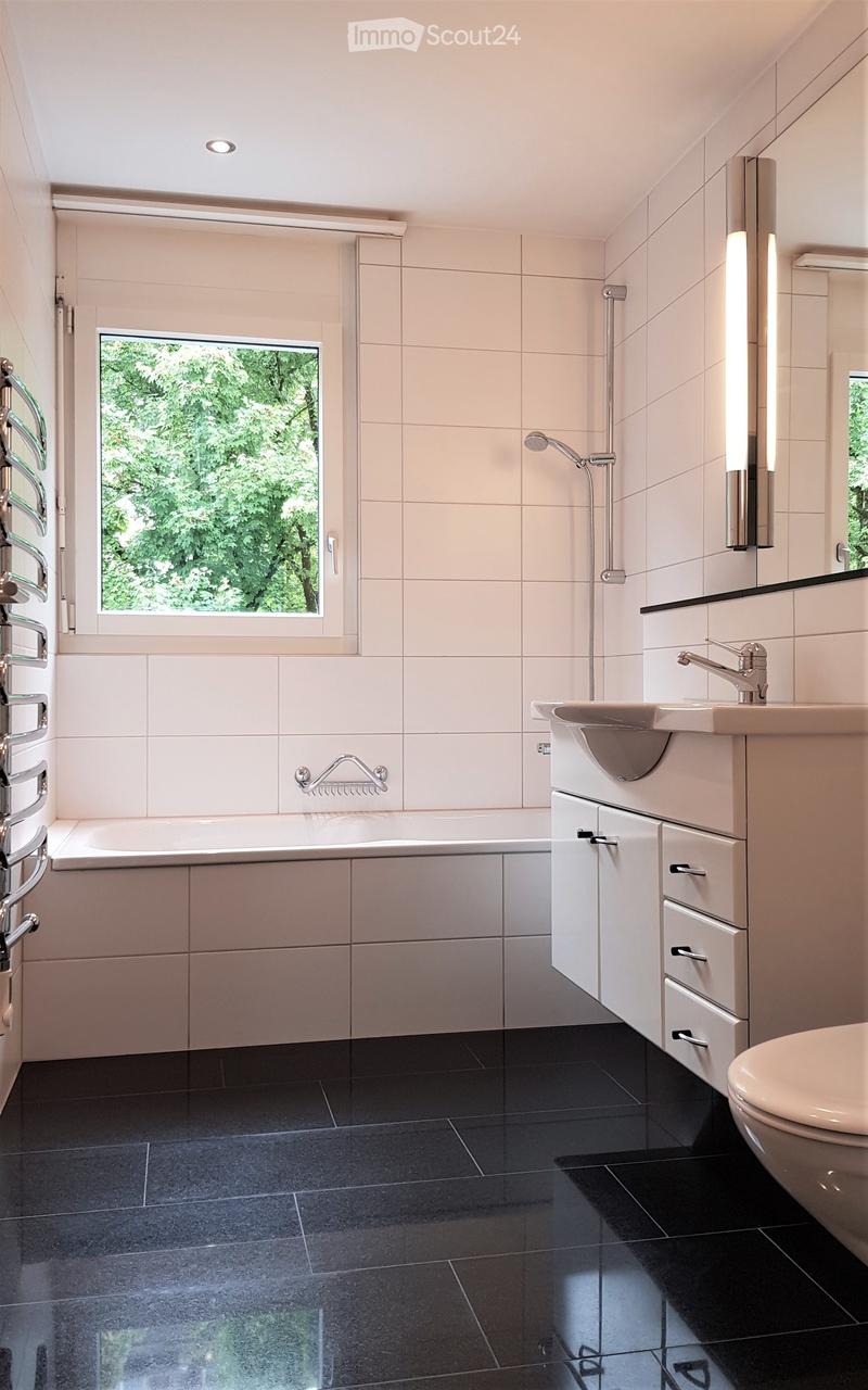 Modern bathroom with a bathtub, sink, toilet, and shower. Features a large window with a view of greenery, white tiles, and dark flooring.