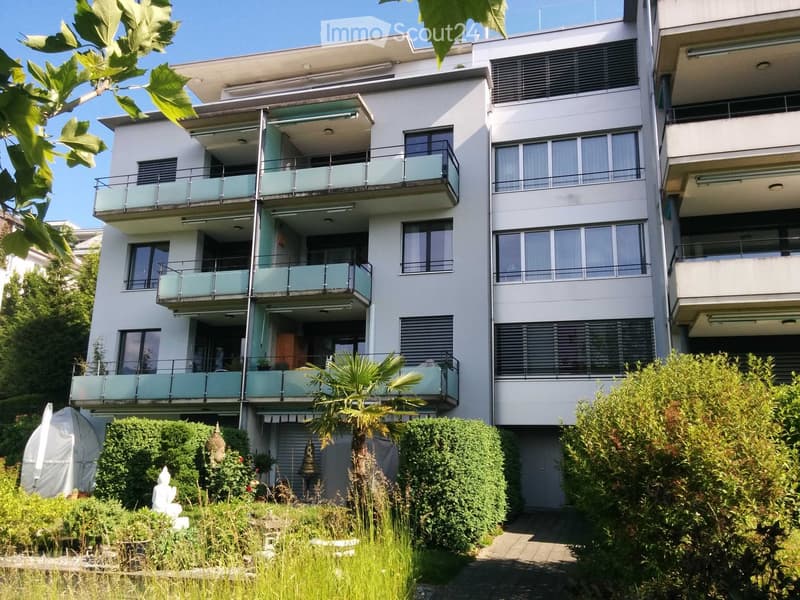 Modern multi-story building with balconies, large windows, and surrounding greenery including a palm tree and a small garden area.