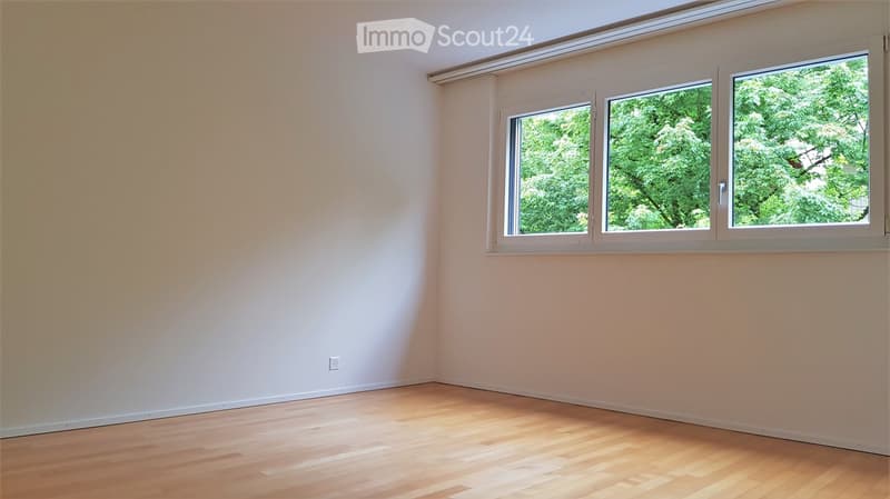 Empty room with light-colored walls and wooden flooring, featuring large windows with white frames and a view of greenery outside.