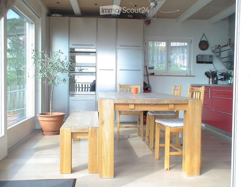 Bright kitchen with a wooden dining table, bench, and chairs, along with modern cabinets, an oven, refrigerator, and microwave. A potted plant adds a touch of greenery.