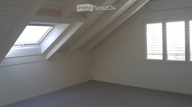 Empty attic room with exposed wooden beams, skylight window, and large window with blinds.