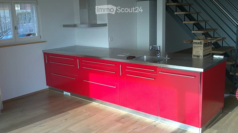 Modern kitchen with red cabinets, stainless steel countertop, sink, and faucet. Wooden floor and window visible.