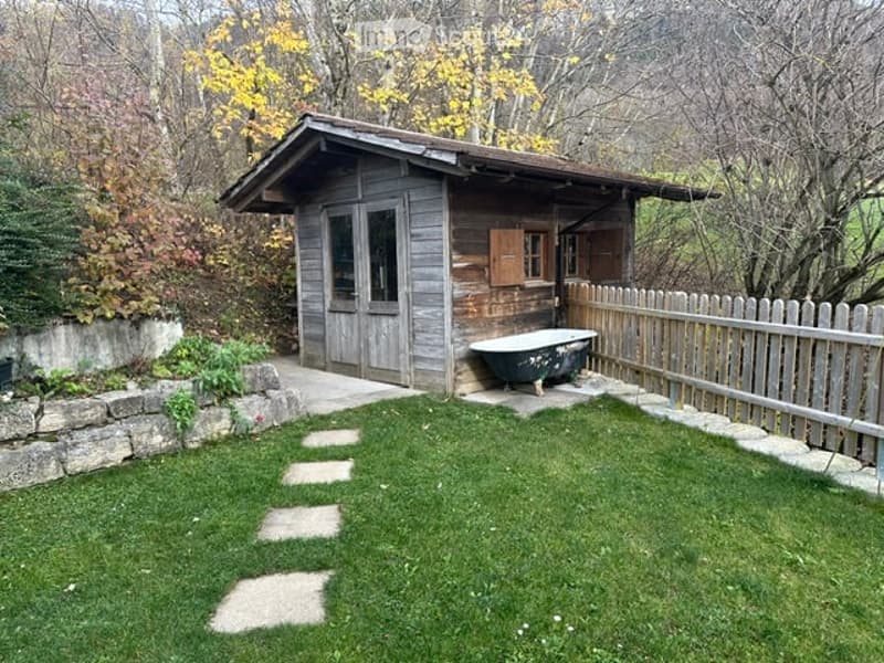 A small wooden cabin with a stone foundation, surrounded by a grassy area with stepping stones leading to the entrance. A wooden fence encloses the space, and a bathtub is placed near the cabin.