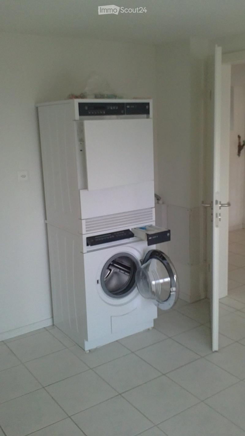 A small bathroom with a stacked washing machine and dishwasher unit, tiled floor, and an open door leading to another room.