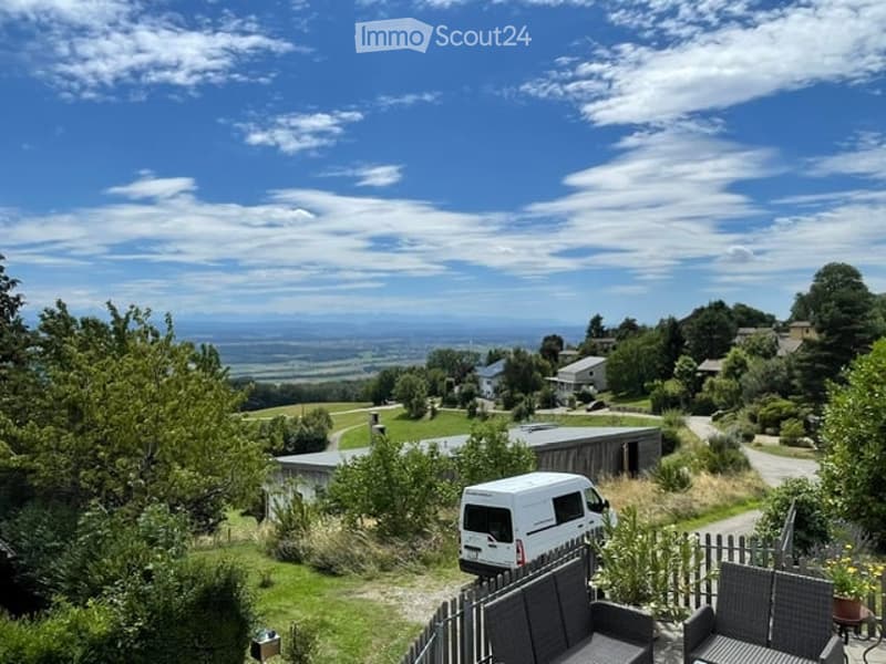Scenic outdoor view with a van, patio furniture, and a panoramic landscape of fields and houses under a clear blue sky.