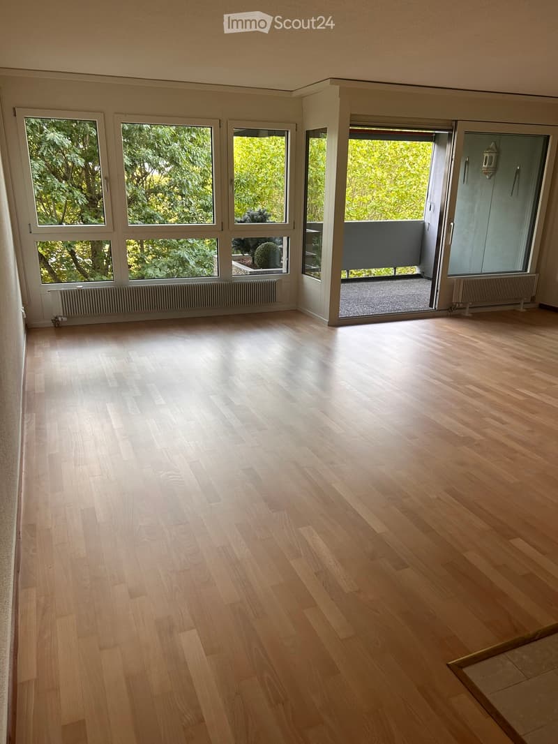 Empty room with light wooden flooring, large windows, and a sliding glass door leading to a balcony.