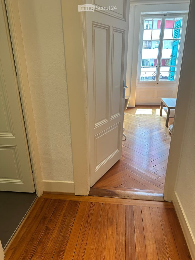A narrow hallway with wooden flooring and white walls, leading to an open doorway that reveals a room with a small table and a window with a view of a balcony.